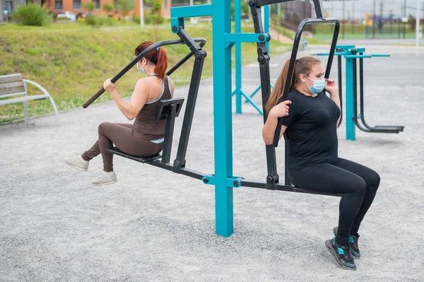 Two Caucasian women in medical masks are exercising on the gym equipment of the sports field. Friends doing fitness outdoors during the spread of the coronavirus.