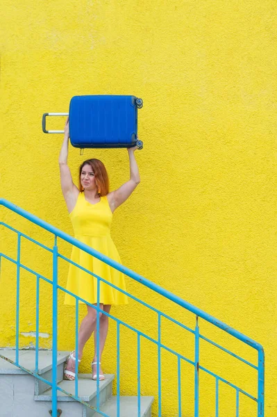 Šťastná zrzavá žena ve žlutých šatech sestupuje ze schodů a radostně houpe modrým kufříkem. Dívka se připravuje na cestu. Koncept letních prázdnin. — Stock fotografie