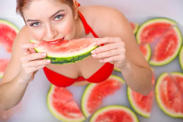 Porträt einer Frau im roten Badeanzug, die eine Wassermelone isst und grinst. Rotschopf Mädchen mit rotem Lippenstift nimmt ein Bad mit Milch und Früchten, um ihre Haut zu verjüngen. Körperpflege. — Stockfoto