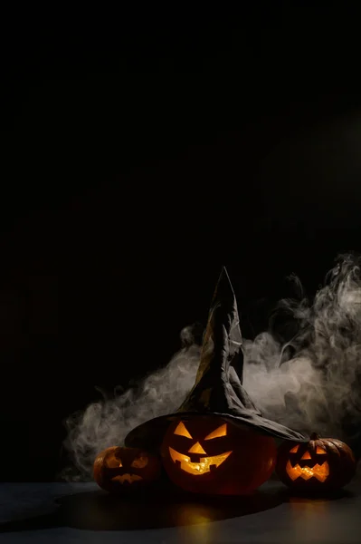 Vertikální Halloweenská karta. Čarodějnický klobouk na dýni s vyřezávanými úšklebky na černém pozadí v mlze. Jack-o-lucerna svítí zevnitř a vypouští kouř. — Stock fotografie