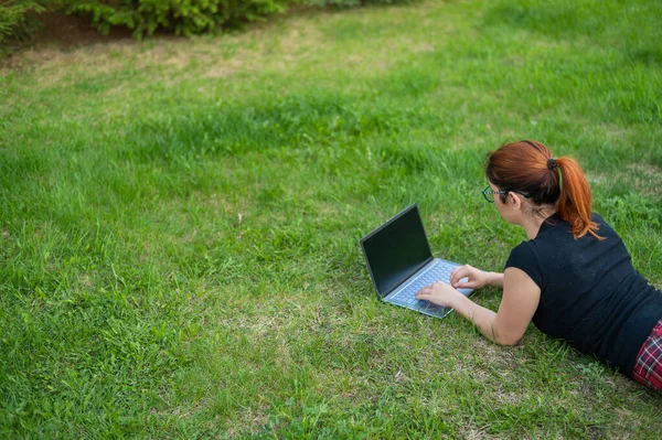 Jovem ruivo mulher remotamente trabalha fora deitado na grama verde. Freelancer menina impressões em um laptop ao ar livre . — Fotografia de Stock