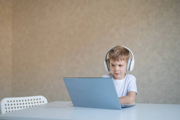 Lindo chico diligente está escribiendo en el teclado y está mirando cuidadosamente a la pantalla del ordenador portátil. El colegial caucásico se sienta en una mesa, escucha música en los auriculares y hace los deberes. Educación a distancia . — Foto de Stock
