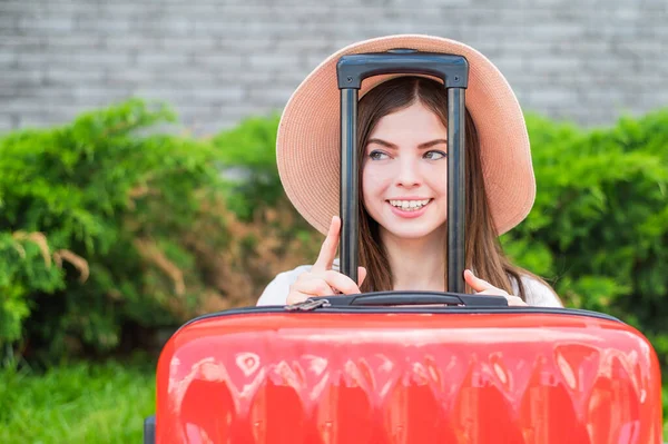 Jonge mooie blanke vrouw met een hoed kijkt uit een rode koffer met een handvat. Gelukkig meisje gaat op reis met bagage. Zomervakantie concept. — Stockfoto