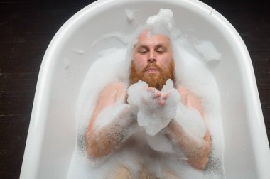 Kızıl sakallı kel bir adamın köpük banyosuna sıçrayan görüntüsü. Komik bir fotoğraf. Çekici kızların parodisi..