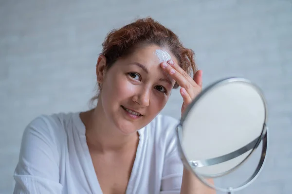 Портрет белой женщины с витилиго использует солнцезащитный крем. Девушка с белым пигментным пятном на лбу смотрит в зеркало и размазана кремом. — стоковое фото
