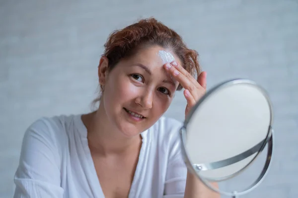 Retrato de una mujer caucásica con vitíligo utiliza protector solar. Una chica con una mancha de pigmento blanco en la frente se mira en el espejo y se unta con crema. — Foto de Stock