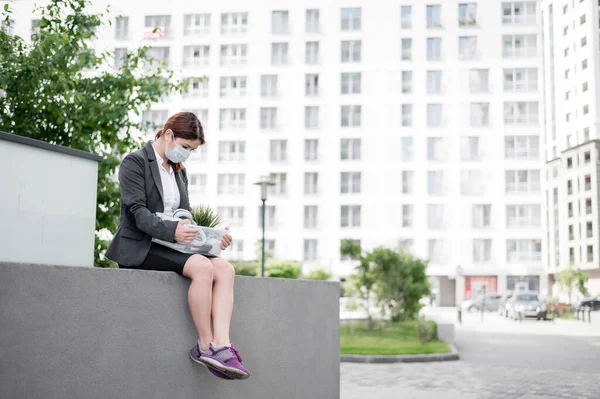 Depressed woman in medical mask sits on parapet outdoors with a box of personal items from the desktop. Unemployed girl alone on the street. Bankruptcy of a company due to quarantine in coronavirus.