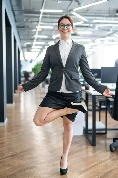 A pacified business woman doing yoga in an open space office. A red-haired smiling female employee in a casual suit and glasses stands in a tree pose at the workplace. Meditation and relaxation.