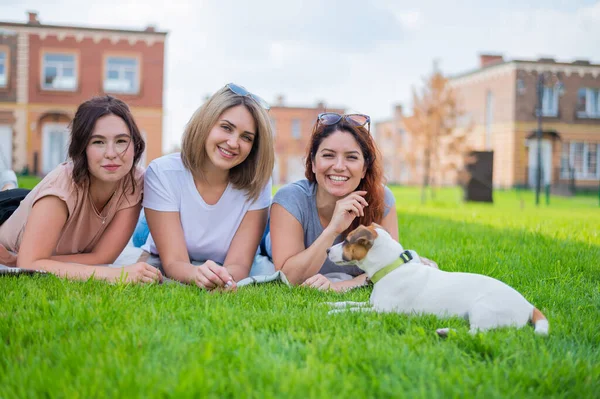 Drie mooie vriendinnen liggen op het gazon op het platteland. Blanke vrouwen en hond op een picknick buiten. blond brunette en roodharige poseren op het groene gras. — Stockfoto