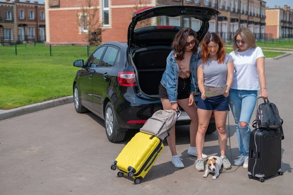 Drei Freunde machen einen Roadtrip in den Sommerferien und schauen auf die Landkarte. Junge Frauen laden Koffer in den Kofferraum eines Autos. Die Mädchen werden sich mit dem Hund ausruhen. — Stockfoto