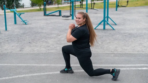 En vacker fet tjej i svart träningsoverall är engagerad i fitness på idrottsplatsen. Ung kvinna lungor utomhus på en varm sommardag. hälsosam livsstil och viktminskning. — Stockfoto