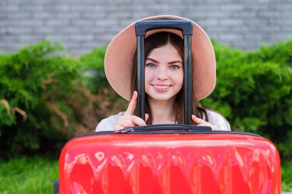 Joven hermosa mujer caucásica en un sombrero se ve fuera de una maleta roja con un mango. La chica feliz va de viaje con el equipaje. Concepto vacaciones de verano. — Foto de Stock