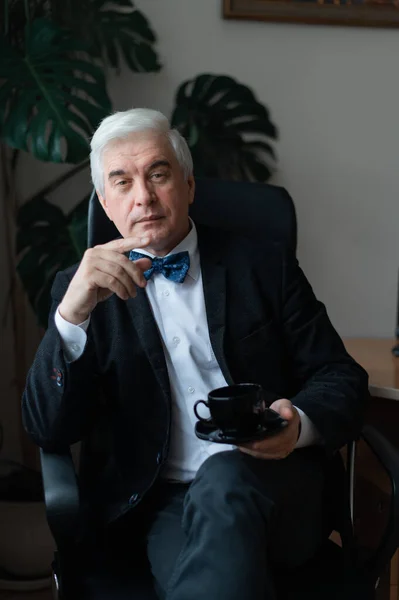 Een aantrekkelijke volwassen man in een pak en een vlinderdas drinkt koffie terwijl hij aan zijn bureau zit. Zelfverzekerde grijze mannelijke baas op een pauze in het kantoor. — Stockfoto