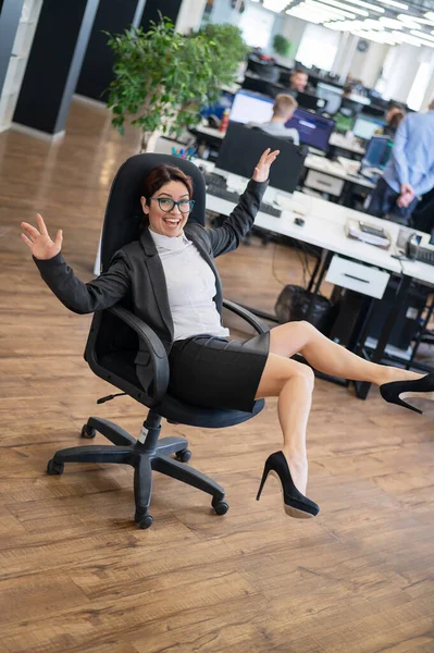 Mujer alegre en un traje monta en una silla de oficina en el lugar de trabajo mientras sus colegas están trabajando. Sonriente dama de negocios divirtiéndose durante el descanso en el trabajo en la oficina de espacio abierto. —  Fotos de Stock