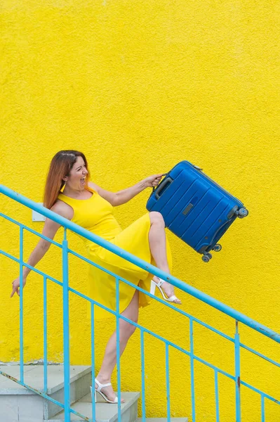 Eine fröhliche rothaarige Frau in einem gelben Kleid läuft die Treppe hinunter und schwingt freudig einen blauen Koffer. Das Mädchen bereitet sich auf die Reise vor. Sommerferienkonzept. — Stockfoto