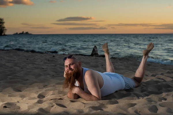 Смешной лысый мужчина с рыжей бородой позирует на пляже на закате. Юмористическая мужская пародия на гламурную девушку. — стоковое фото