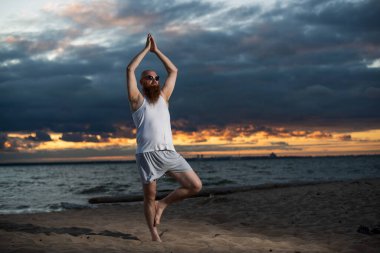 Kızıl sakallı kel bir adam gün batımında plajda yoga yapıyor. Tişörtlü ve güneş gözlüklü komik bir adam deniz kıyısında meditasyon yapıyor..