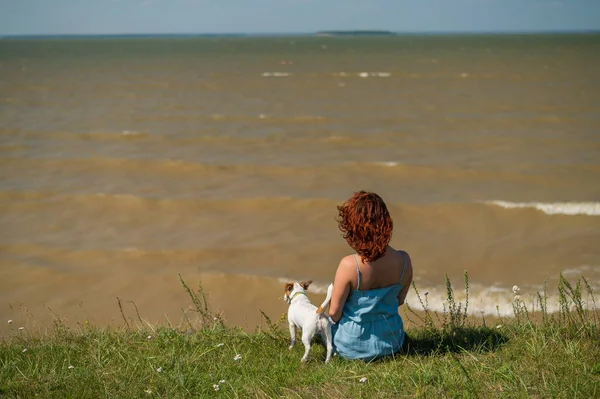 Une femme est assise sur la rive de la rivière avec un chien. Vue arrière d'une fille se reposant dans la nature avec un animal de compagnie. — Photo