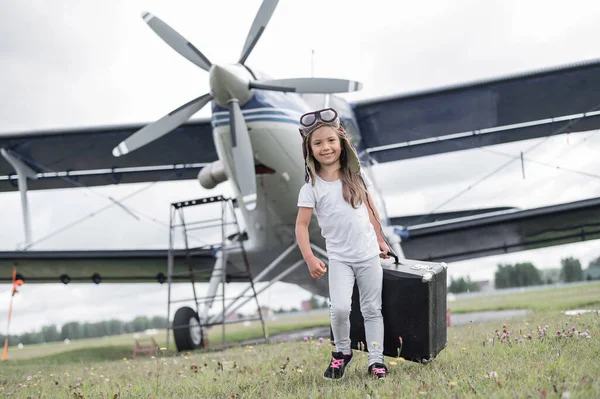 Een klein meisje in een pilotenkostuum draagt een retro koffer en loopt over het vliegveld. Een kind met hoed en bril gaat met het vliegtuig op reis.. — Stockfoto