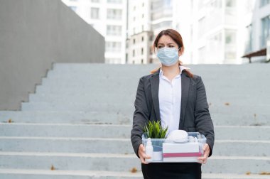 Maskeli mutsuz kadın merdivenlerin arkasında kişisel eşyalarla dolu bir kutuyla cadde boyunca yürüyor. Kadın bir ofis çalışanı kovulmuş. Salgın sırasında ekonomik kriz covid 19.