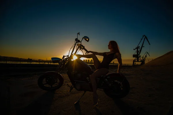 Mujer pelirroja joven en lencería erótica en tacones altos se sienta en una motocicleta. Silueta de una chica sexy en una bicicleta. Paisaje industrial. —  Fotos de Stock