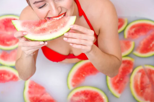 Πορτρέτο μιας γυναίκας με κόκκινο μαγιό που τρώει καρπούζι και χαμογελάει. Redhead κορίτσι με κόκκινο κραγιόν παίρνει ένα μπάνιο με γάλα και φρούτα για να αναζωογονήσει το δέρμα της. Φροντίδα σώματος. — Φωτογραφία Αρχείου