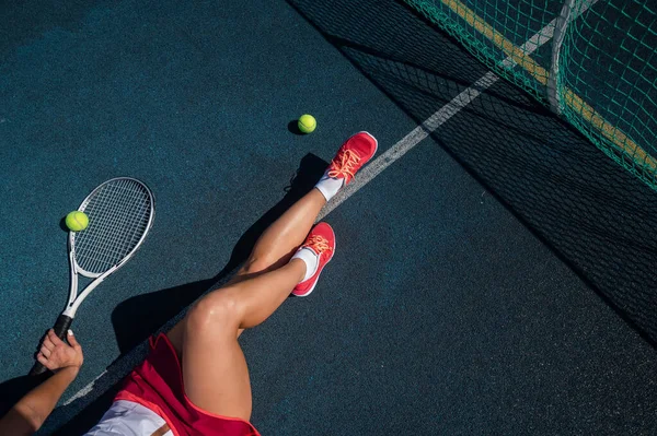 Spor etekli yüzü olmayan bir kız tenis kortunda oturuyor ve elinde bir roket tutuyor. Kadın bacaklarının üst görüntüsü.