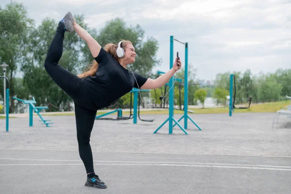 Belle jeune femme grosse en casque fait un patineur artistique et prend un selfie. Une fille souriante potelée écoute de la musique et se réchauffe avant de faire du jogging à l'extérieur. Sports pour perdre du poids. — Photo