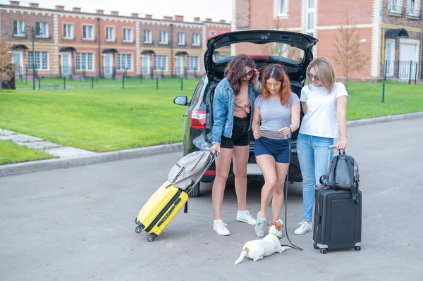 Drei Freunde machen einen Roadtrip in den Sommerferien und schauen auf die Landkarte. Junge Frauen laden Koffer in den Kofferraum eines Autos. Die Mädchen werden sich mit dem Hund ausruhen. — Stockfoto