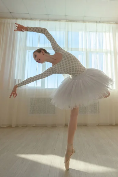 Die schöne anmutige Ballerina übt im Saal vor dem Hintergrund eines Fensters. Schlanke Frau in Tutu und Spitzenschuhen in einem Tanzkurs. — Stockfoto