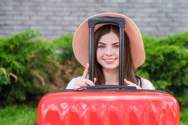 Jonge mooie blanke vrouw met een hoed kijkt uit een rode koffer met een handvat. Gelukkig meisje gaat op reis met bagage. Zomervakantie concept. — Stockfoto