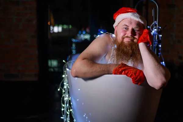 Image humoristique d'un père Noël maléfique prenant un bain avec des lumières de Noël. Un homme avec une barbe rouge dans un chapeau et des sous-vêtements du Père Noël tient une guirlande dans ses mains. — Photo