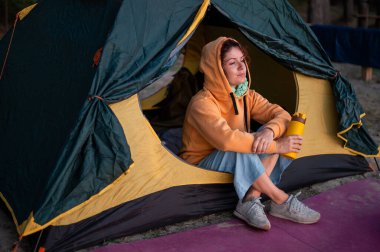 Kafkasyalı sakin bir kadın turist çadırında tek başına oturmuş termostan sıcak çay içiyor. Kız kamp alanındaki termo bardaktan içip gün batımını izlemekten zevk alıyor..