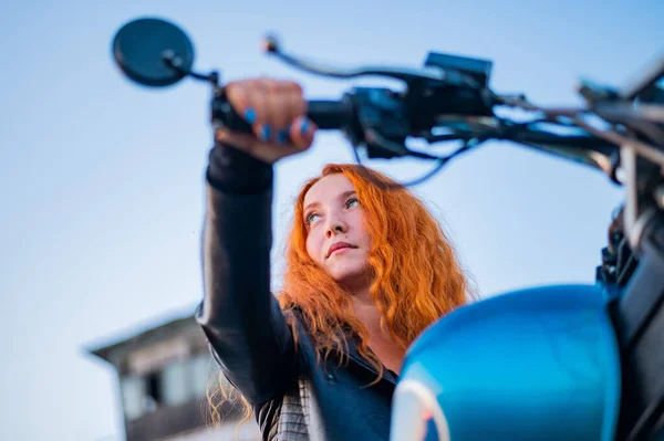 Mulher ruiva encaracolado em um casaco de couro preto senta-se em uma motocicleta. Retrato de uma menina séria dirigindo uma bicicleta. — Fotografia de Stock