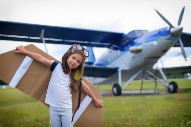 A little girl in a pilots costume with cardboard wings runs on the lawn against the backdrop of the plane. A child in a hat and glasses dreams of flying on an airplane. clipart