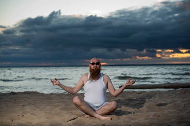 Kızıl sakallı kel bir adam gün batımında plajda yoga yapıyor. Tişörtlü ve güneş gözlüklü komik bir adam deniz kıyısında meditasyon yapıyor..