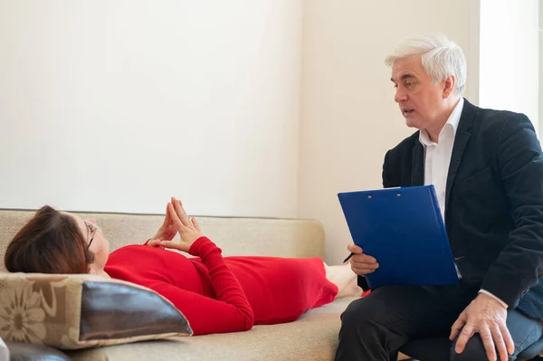 Primer plano de un psicoterapeuta masculino escribiendo a mano en un portapapeles. Una mujer vestida de rojo yace en un sofá y habla de sus problemas a un psicólogo en una sesión. Salud mental. —  Fotos de Stock
