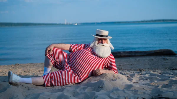 Un uomo anziano in costume da bagno retrò a righe prende il sole sulla spiaggia. Un vecchio uomo barbuto dai capelli grigi con un cappello giace sulla sabbia vicino al mare. — Foto Stock