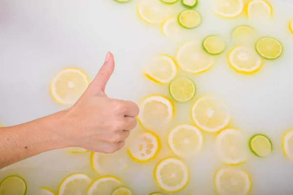 A womans hand shows her thumb against the background of a milk bath with citrus fruits. Woman gestures approval while taking a bath with lemons and limes. — Stock Photo, Image