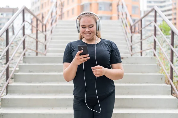 A fat young woman goes down the stairs and listens to music on a smartphone. Obese girl with headphones resting after jogging. — Stock Photo, Image