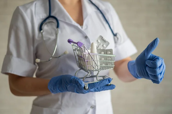 Egy arctalan orvos orvosi kabáttal és sztetoszkóppal tart egy kis bevásárlókocsit tele gyógyszerekkel. Gyógyszerészeti fogalom. A gyógyszerész mutat egy hüvelykujj felfelé. — Stock Fotó