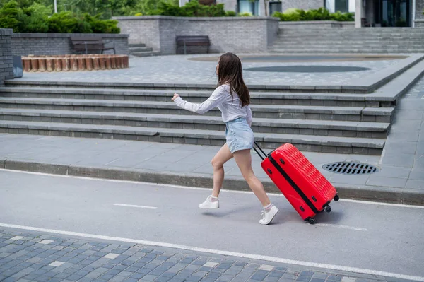 Glücklich schöne kaukasische Frau läuft mit einem großen roten Koffer nach draußen. Aufgeregtes Mädchen kommt zu spät ins Flugzeug. — Stockfoto