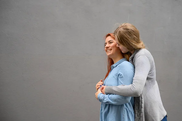 Twee vrolijke vrouwen knuffelend tegen de achtergrond van een grijze muur. Zachte knuffels van een lesbisch echtpaar. LGBT huwelijk van hetzelfde geslacht. — Stockfoto