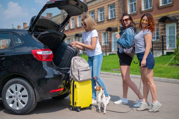 Drei Freundinnen gehen auf Roadtrip. Frauen laden ihr Gepäck in den Kofferraum eines Autos. Mädchen fahren mit dem Hund in den Urlaub. — Stockfoto