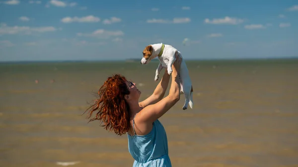 Счастливая женщина обнимается с собакой у моря. Рыжая белая девушка держит щенка Джека Рассела Терьера. Любовь между домашним животным и владельцем. — стоковое фото