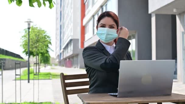 Happy business žena odstraňuje lékařskou masku při práci na notebooku na letní terase. Usmívající se dívka v obleku se těší svobodě, zatímco sedí v kavárně. Konec karantény. — Stock video