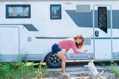 Güzel beyaz kadın karavanın yanında bir köpeği eğitiyor. Hayvanınla seyyar bir karavanla seyahat et. Kızıl saçlı kız Jack Russell Terrier ile oynuyor. Kadın karavan sürücüsü.