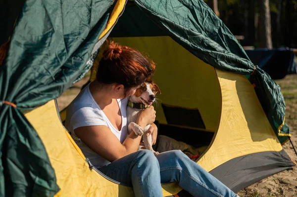 Piękna rudowłosa kobieta odpoczywa w naturze ze swoim zwierzakiem. Dziewczyna siedzi w namiocie turystycznym i przytula w lesie Jacka Russella Terriera. Pies i jego właścicielka na kempingu. — Zdjęcie stockowe