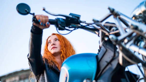 Mulher ruiva encaracolado em um casaco de couro preto senta-se em uma motocicleta. Retrato de uma menina séria dirigindo uma bicicleta. — Fotografia de Stock