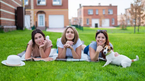 Drei schöne Freundinnen ruhen sich auf dem Rasen im Grünen aus. Kaukasische Frauen und Hund bei einem Picknick im Freien. Blonde Brünette und Rotschopf posieren auf dem grünen Gras. — Stockfoto